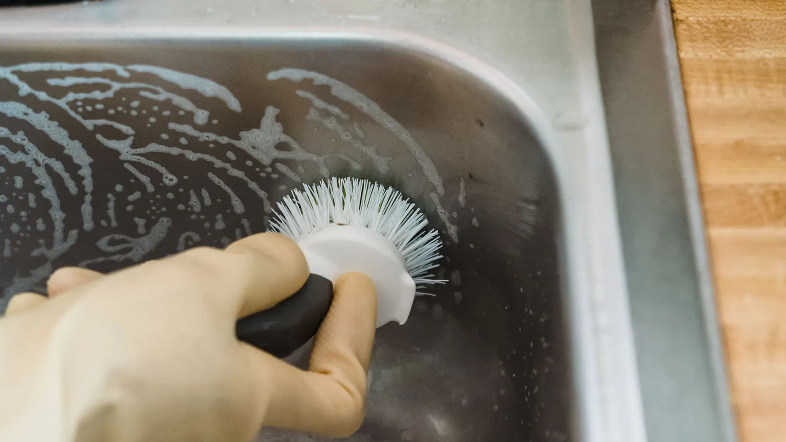 Gloved hand using a round-bristle brush in a soapy metal sink basin.