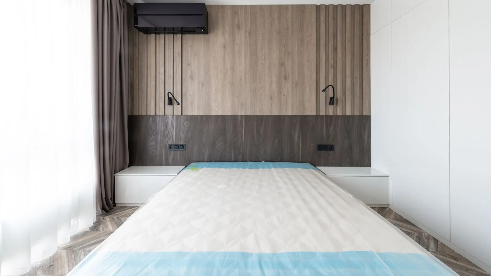 Mattress with protective wrapping on a bed frame in a bright modern bedroom.
