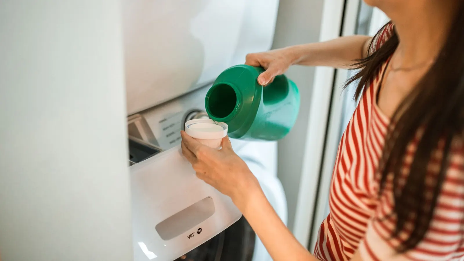 Person measuring liquid detergent beside an open washing-machine dispenser.